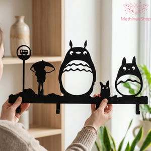 Könnte beinhalten: Schwarzer Metall-Wandhaken mit Silhouetten von Figuren aus einem beliebten Zeichentrickfilm. Das Design umfasst ein Bushaltestellenschild, eine Figur mit einem Regenschirm und zwei große, runde Charaktere. Der Haken hat drei Haken zum Aufhängen von Gegenständen.