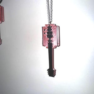 May include: A pink and black pendant necklace with a razor blade design. The pendant is suspended from a silver chain. The razor blade design has a pink body with black detailing. The necklace is set against a white backdrop.