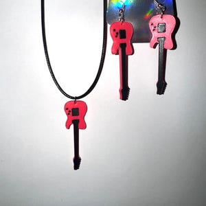 May include: A necklace and earrings set with guitar-shaped pendants. The pendants are pink with black detailing. The necklace has a black cord, and the earrings have silver hooks. The set is presented against a white backdrop.