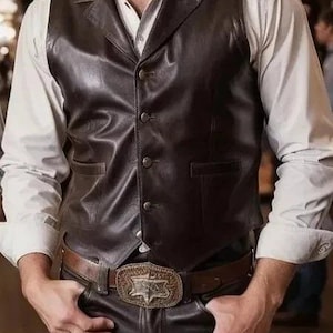 May include: A dark brown leather vest with a notched collar and button closure. The vest is worn over a white button-down shirt. The outfit includes a brown leather belt with a decorative buckle and dark brown leather pants. The overall style is Western-inspired.
