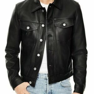 Può includere: Giacca di pelle nera dal design classico. La giacca presenta un colletto, due tasche sul petto e bottoni argentati. La giacca è indossata sopra una camicia bianca e jeans blu.