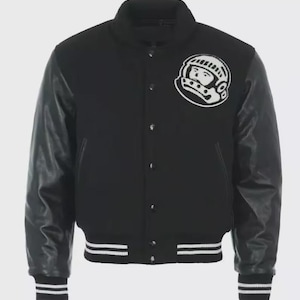 May include: Black varsity jacket with a white astronaut patch on the chest. The jacket features black leather sleeves, a black body, and white striped cuffs and hem. A classic, stylish outerwear piece.