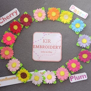 Puede incluir: Una colección de aplicaciones de flores de fieltro en varios colores, incluyendo rojo, rosa, amarillo, naranja, azul, morado y verde. Las flores están dispuestas en un círculo alrededor de una tarjeta blanca con el texto "KIR EMBROIDERY only on Etsy".