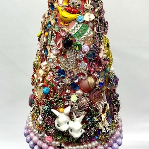 Op de afbeelding: Een decoratieve paasboom, versierd met een veelheid aan kleurrijke juwelen, kralen en bedels. De boom heeft een roze basis en is versierd met paasversieringen, waaronder een gele Peep, eieren en witte konijnenbeeldjes. De top van de boom is versierd met roze en gouden versieringen.