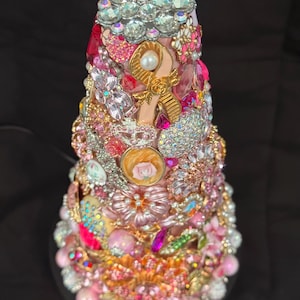 May include: A decorative, cone-shaped object covered in an array of pink, gold, and clear jewels. The embellishments include rhinestones, pearls, and floral accents. A gold ribbon is also featured. The base is adorned with pink and white pearl-like beads.