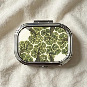 Peut inclure: Un pilulier rectangulaire en métal aux coins arrondis avec une finition argentée polie. Le couvercle présente un motif graphique répétitif d'arbres verts stylisés avec des troncs brun foncé et un feuillage texturé sur fond crème.