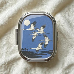 Peut inclure: Un pilulier rectangulaire en métal argenté aux coins arrondis avec un insert supérieur brillant. L'insert présente un ciel bleu avec une lune blanche et cinq grues blanches en vol au-dessus d'un paysage sombre avec des silhouettes d'arbres et d'eau.