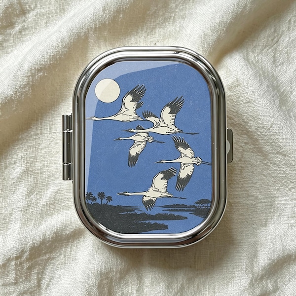 May include: A rectangular silver metal pill case with rounded corners featuring a glossy top insert. The insert displays a blue sky with a white moon and five white cranes in flight above a dark landscape with silhouettes of trees and water.