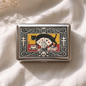 Pode incluir: Uma caixa retangular de metal prateado com uma ilustração gráfica de um gato preto, uma mão segurando cartas de jogar, uma xícara de chá e uma caveira. O design inclui uma ampulheta, dois símbolos de cruz e bordas florais em fundo preto.