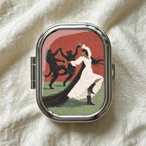 Pode incluir: Um porta-comprimidos retangular de metal prateado com uma ilustração de uma pessoa de cabelos longos e escuros, vestindo um vestido branco e botas pretas, dançando com dois gatos pretos sobre um fundo de sol vermelho e relva verde.