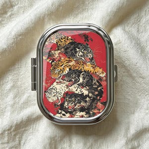 Puede incluir: Un pastillero rectangular de metal con esquinas redondeadas y un acabado plateado pulido. La tapa presenta un fondo rojo vibrante decorado con una ilustración detallada de varios tigres en diferentes colores, incluyendo negro, blanco y naranja.