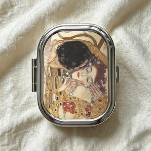Peut inclure: Un pilulier rectangulaire en métal aux coins arrondis avec une finition argentée polie. Le couvercle présente une reproduction imprimée du Baiser de Gustav Klimt, montrant deux personnages enlacés avec des motifs dorés, bruns et floraux.