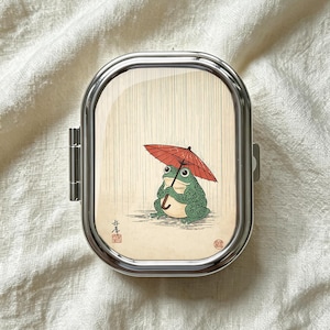 Pode incluir: Uma caixa de comprimidos retangular de metal com cantos arredondados, apresentando uma ilustração impressa de um sapo verde segurando um guarda-chuva vermelho na chuva. A arte inclui linhas verticais sutis para a chuva e dois pequenos selos vermelhos.