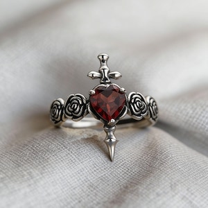 Op de afbeelding: Een zilveren ring met een hartvormige, dieprode edelsteen in het midden, doorboord door een dolk. De ring is versierd met rozen en geplaatst tegen een zachte, neutrale achtergrond. Het ontwerp heeft een gotisch of romantisch thema.