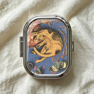 May include: A rectangular silver metal pill case with rounded corners featuring a colorful illustration of a sleeping orange cat curled up on a blue surface with orange yarn, small fish, and open metal cans scattered around the feline figure.