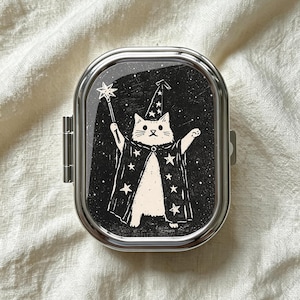 May include: A rectangular silver metal pill case with rounded corners featuring a black graphic print of a white cat dressed as a wizard. The cat wears a star-patterned robe and hat while holding a star-tipped wand against a starry night sky.