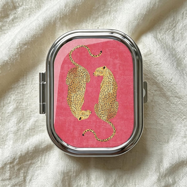 May include: A rectangular silver metal pill case with rounded corners featuring a vibrant pink background and a graphic illustration of two spotted leopards facing each other. The case has a hinged side closure and a polished metallic finish.
