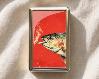 以下が含まれることがあります： 口に火のついたタバコをくわえた魚のグラフィックイラストが描かれた、長方形の銀色の金属製シガレットケース。イラストの背景は鮮やかな赤色で、魚の頭の後ろには明るいピンク色の円形の形が配置されています。