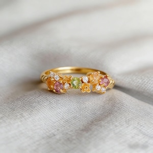 Könnte beinhalten: Ein goldener Ring mit einer Ansammlung farbiger Edelsteine. Der Ring hat rosa, orange, grüne und weiße Steine. Der Ring ist auf einem goldenen Band mit einem strukturierten Muster angebracht.