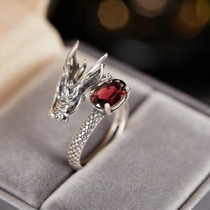 May include: A silver dragon ring featuring a red oval gemstone. The dragon's head design wraps around the finger, with detailed scales and wings. The ring is presented in a grey velvet box.