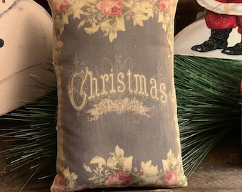 Puede incluir: Una almohada gris y blanca con un patrón floral y la palabra "Christmas" en una fuente decorativa. La almohada está sentada sobre una superficie de madera.