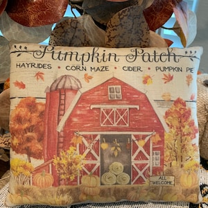 May include: A decorative pillow with a fall theme. The pillow features a red barn with a white door and windows, hay bales, pumpkins, and fall foliage. The text "Pumpkin Patch" is written in a decorative font above the barn. The text "Hayrides, Corn Maze, Cider, Pumpkin Pie" is written below the barn. The text "All Welcome" is written on a sign in front of the barn.