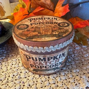 May include: A round, brown cardboard container labeled Pumpkin Patch Popcorn with a vintage-style graphic of pumpkins and a ghost. The lid and sides feature decorative text and a rustic jute trim border around the top edge of the container.