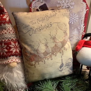 May include: A beige pillow with a vintage design featuring a scene of Santa Claus in a sleigh pulled by reindeer. The pillow has the text "'Twas The Night Before Christmas" printed on it.