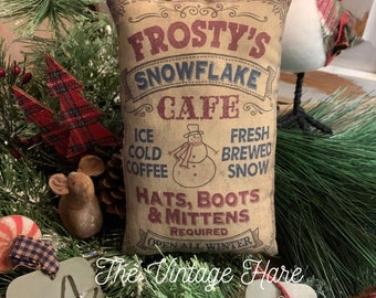 May include: A brown fabric pillow with the text "Frosty's Snowflake Cafe" in red and blue lettering. The pillow also has the text "Ice Cold Coffee", "Fresh Brewed Snow", "Hats, Boots & Mittens Required", and "Open All Winter".
