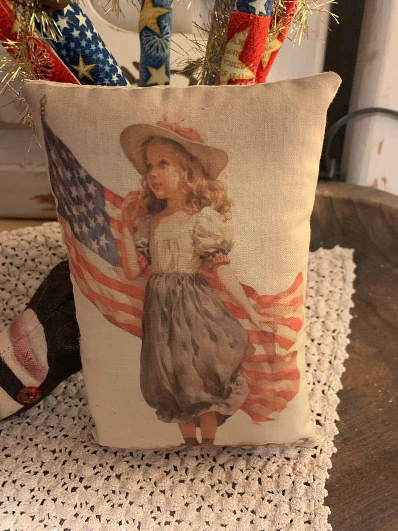 May include: A white pillow with a vintage illustration of a young girl holding an American flag. The girl is wearing a white dress and a straw hat with a red ribbon.