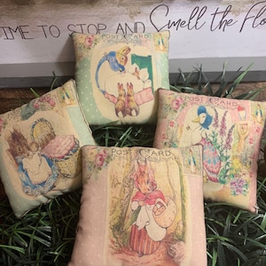 May include: Four small, square decorative pillows with vintage-style illustrations of rabbits and ducks. Each pillow features a postcard design with floral accents and pastel colors. The pillows are arranged on a bed of green grass.