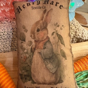 May include: A tan pillow with a vintage design featuring a brown rabbit with a blue vest and the text "Henry Hare Textile Co. - Loom Woven Fabrics - Family Owned Since 1893".