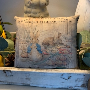 May include: A decorative pillow with a vintage design featuring two rabbits and the text "Peter Rabbit Farms Fresh Vegetables".