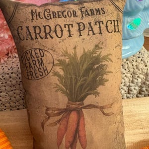 May include: A brown and tan pillow with a vintage design featuring a bunch of carrots with green tops and the text "McGregor Farms Carrot Patch" and "Picked Farm Fresh" in a circle. The pillow also has the text "Family Owned - Open Daily" at the bottom.