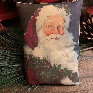 May include: A rectangular pillow featuring a vintage illustration of Santa Claus. The image shows Santa with a white beard and red hat, surrounded by holly and berries. The pillow has a dark blue background and is placed on a wooden surface.