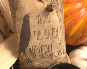 Può includere: Un cuscino decorativo dall'estetica vintage. Presenta una silhouette di corvo e il testo "Quoth the Raven Nevermore" all'interno di una cornice ornata. Una piccola campana è attaccata con una spilla da balia. L'immagine include mais, zucche e castagne.
