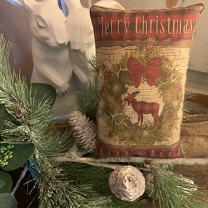 Könnte beinhalten: Ein dekoratives Kissen mit einem rot-grün-braunen Design, das ein Elch und einen Kranz mit einer roten Schleife zeigt. Das Kissen hat den Text "Merry Christmas" auf der Vorderseite.