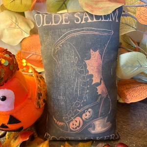 May include: A decorative pillow with a vintage-style design featuring a witch's boot, pumpkins, and the text "OLDE SALEM BOOT MAKERS". The pillow is dark with orange and cream accents, perfect for Halloween decor.