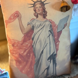 May include: A cushion with a vintage print of the Statue of Liberty. The image depicts Lady Liberty in a flowing gown, holding a tablet and a star-spangled drape. A small, aged brass bell is attached to the cushion.
