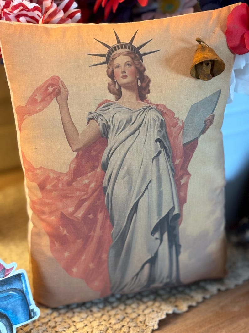 May include: A cushion with a vintage print of the Statue of Liberty. The image depicts Lady Liberty in a flowing gown, holding a tablet and a star-spangled drape. A small, aged brass bell is attached to the cushion.