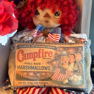 May include: A handmade doll with red yarn hair and a patriotic bow tie, holding a pillow-shaped sign. The sign reads "Campfire Brand Marshmallows" and features a vintage illustration of children celebrating with fireworks. Net weight is 12 oz.