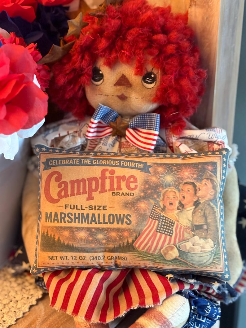 May include: A handmade doll with red yarn hair and a patriotic bow tie, holding a pillow-shaped sign. The sign reads "Campfire Brand Marshmallows" and features a vintage illustration of children celebrating with fireworks. Net weight is 12 oz.