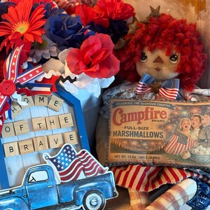 May include: A patriotic display featuring a blue pickup truck carrying an American flag, a doll with red yarn hair, and a vintage Campfire Marshmallows box. Red, white, and blue flowers and a wooden sign that reads "Home of the Brave" complete the arrangement.