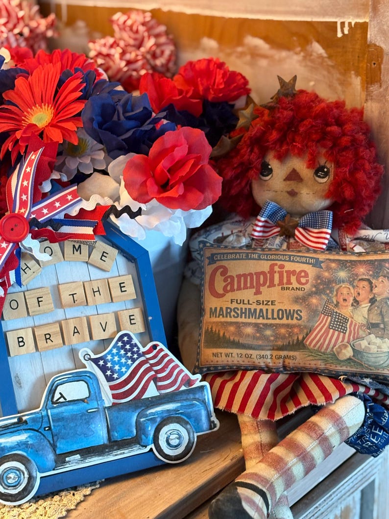 May include: A patriotic display featuring a blue pickup truck carrying an American flag, a doll with red yarn hair, and a vintage Campfire Marshmallows box. Red, white, and blue flowers and a wooden sign that reads "Home of the Brave" complete the arrangement.
