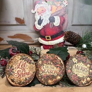 Könnte beinhalten: Drei kreisförmige Holzornamente mit festlichen Weihnachtsmotiven. Das linke Ornament trägt die Aufschrift Let It Snow, das mittlere Tis The Season und das rechte Merry Christmas. Dahinter steht ein Weihnachtsmann-Aufsteller.