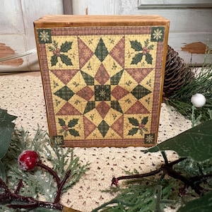 Könnte beinhalten: Ein rechteckiger Holzblock mit einem aufgedruckten Quilt-Muster, das einen zentralen Stern, geometrische Formen in Grün-, Rot- und Gelbtönen sowie Eckakzente aus Stechpalmenblättern und roten Beeren auf hellem Grund zeigt.