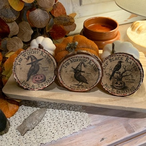 May include: Three round wooden coasters with black and white illustrations of ravens, pumpkins, and trees. The coasters feature the text "The Witchy Raven Inn & Tavern", "The Mischief Raven Inn & Pub", and "Ole' Black Raven Bed & Breakfast".