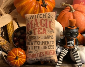 May include: A decorative pillow with a vintage design featuring a witch's magic tea that cures curses, charms, bewitchments, evil eye, and jinxes. The pillow is surrounded by pumpkins and corn. The text on the pillow reads "Witch's Magic Tea Cures Curses Charms Bewitchments Evil Eye Jinxes Also Eliminates Warts Purchase All Potion Shops."