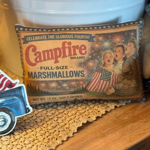 May include: A vintage-style pillow featuring a marshmallow bag design. The bag reads "Campfire Brand Full-Size Marshmallows" with the text "Celebrate the Glorious Fourth!" and a picture of children with American flags. Net weight is 12 oz.