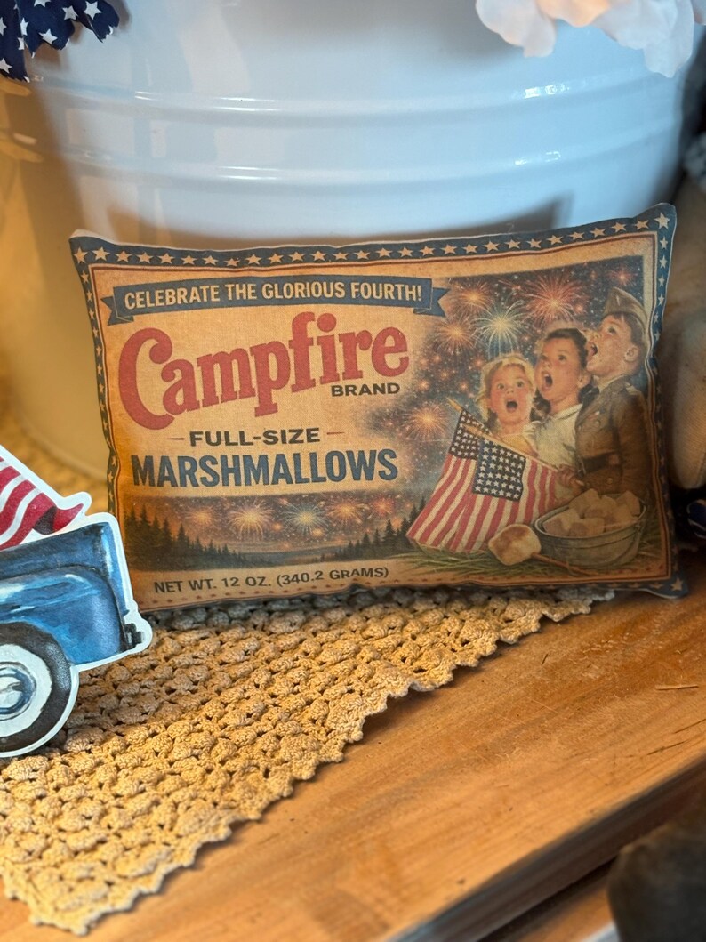 May include: A vintage-style pillow featuring a marshmallow bag design. The bag reads "Campfire Brand Full-Size Marshmallows" with the text "Celebrate the Glorious Fourth!" and a picture of children with American flags. Net weight is 12 oz.